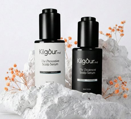 Front view of two KilgourMD scalp serums, white Prevention and black Treatment, 50 ml, on white rocks with orange flowers