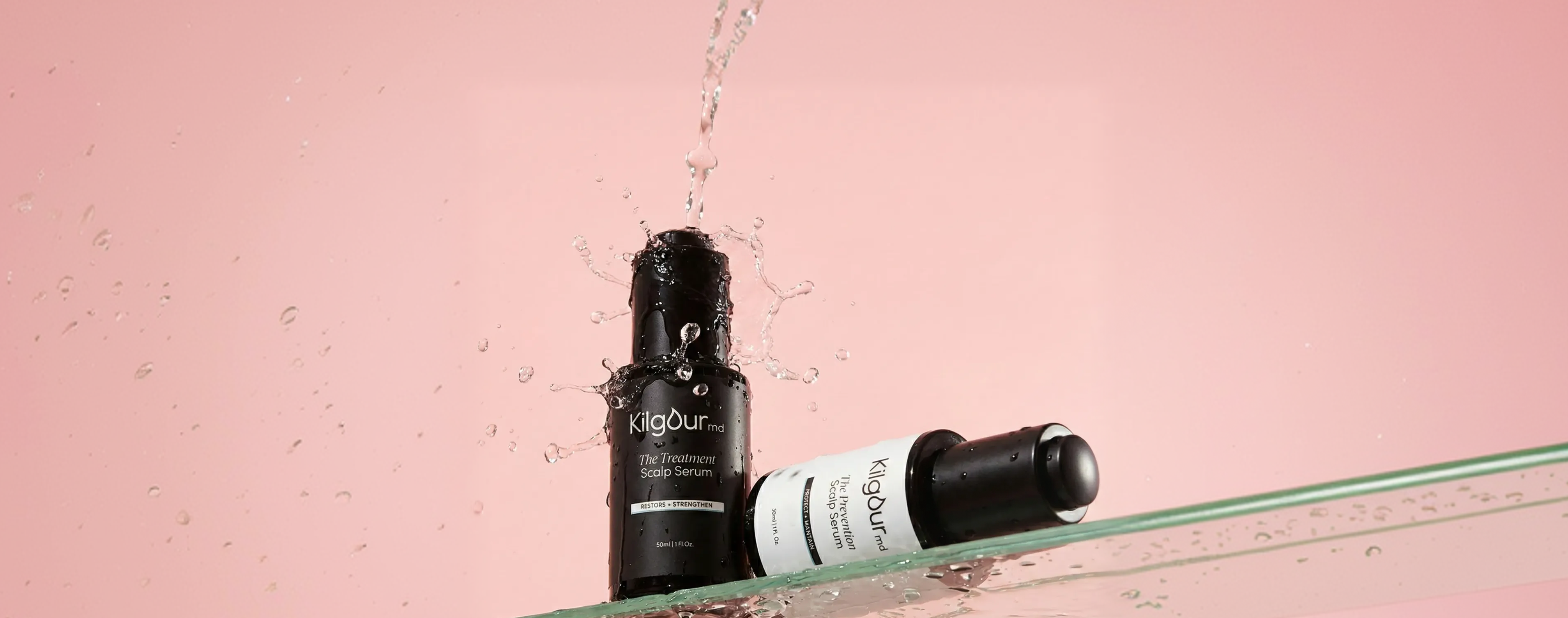 Two KilgourMD scalp serums, one upright splashed with water and one lying on a glass shelf against a pink background