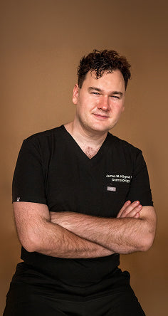 Dr. James Kilgour posing with crossed arms in black scrubs against a brown background