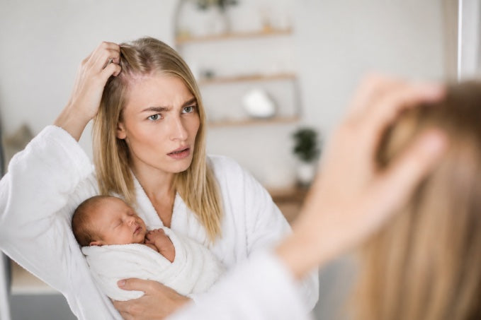 Postpartum mother holding a sleeping baby while checking her hair in a mirror