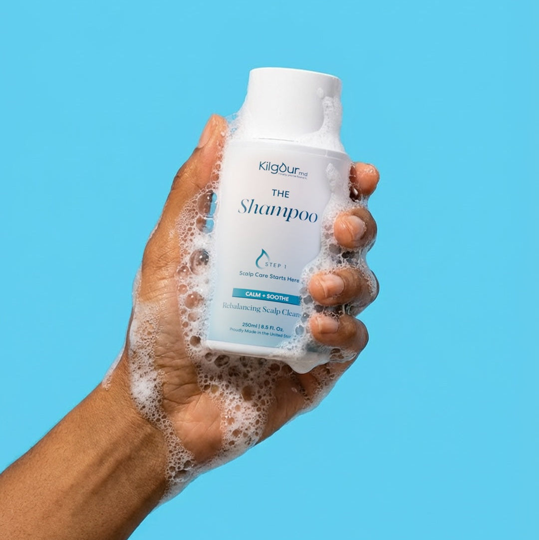 Close-up of KilgourMD The Shampoo (Calm + Soothe) bottle held in a soapy hand, showcasing the front label and lather against a blue background